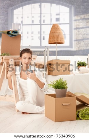 Portrait of young attractive female moving to new house, sitting on floor, drinking tea, boxes around.?