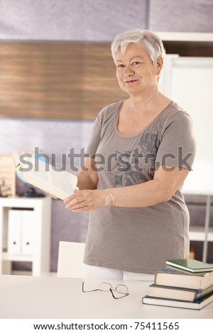 Portrait of senior teacher standing at desk in classroom, holding textbook, looking at camera.?