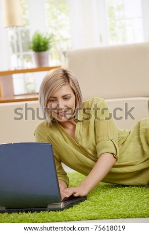 Smiling woman typing on laptop keyboard, lying on floor at home.?