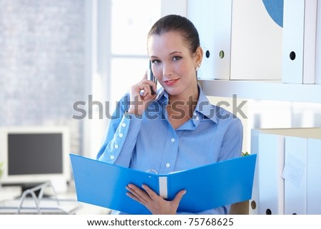 Young smiling attractive assistant on mobile phone call, holding folder in office, smiling at camera.