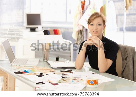 Portrait of attractive female fashion designer sitting at office desk, smiling.