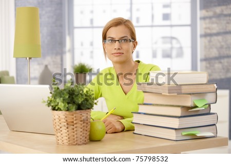 Young woman studying for university at home, sitting at desk with books and laptop computer, smiling at camera.?