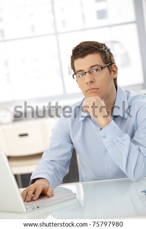 Portrait of thinking businessman sitting in office with laptop computer, looking at camera.?