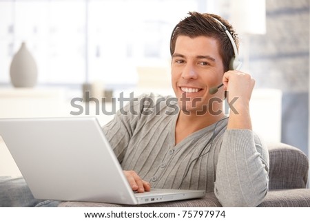 Happy guy using headset and laptop computer, chatting on Internet, smiling at camera.?