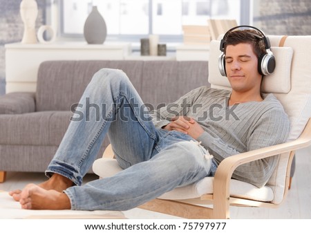 Handsome guy enjoying music on headphones, sitting in armchair with eyes closed, smiling.?