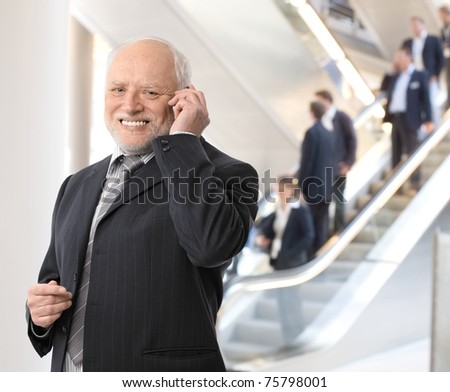 Happy senior businessman standing in office lobby, taking phone call, smiling at camera.?