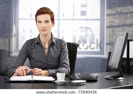 Determined businesswoman sitting at desk, working with personal organizer, looking at camera.?