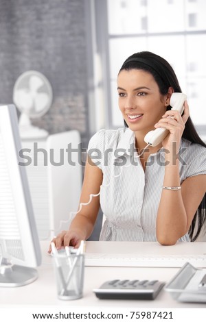 Happy office girl at desk working on desktop computer, using landline phone, smiling.?