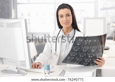 Portrait of doctor sitting in office holding xray image, working on computer, smiling at camera.?