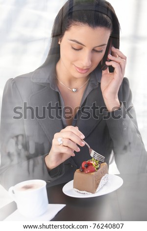 Smart office worker woman on phone call, sitting in cafe, having chocolate cake and coffee, smiling, picture through window.?