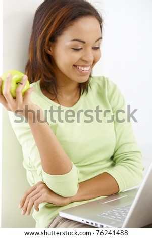 Happy girl having green apple and looking at laptop computer screen, laughing.?