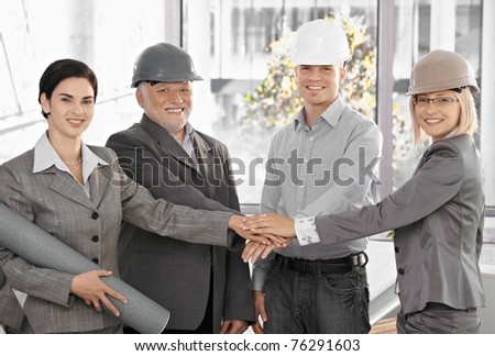 Architect team in office holding hands in unity, expressing teamwork, wearing hardhat, smiling at camera.?