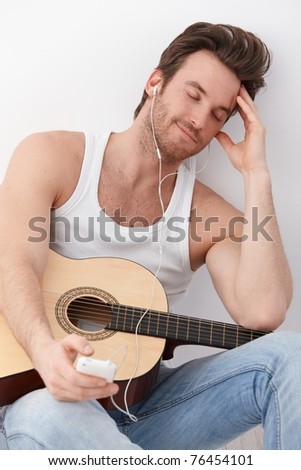 Sexy guitar player sitting on floor, listening to music through headphones, having mp3 player, smiling.