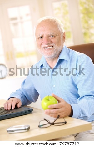Active senior man sitting at desk at home, smiling, holding apple.?