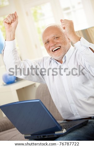 Old man celebrating at home, laughing and raising arms, having laptop computer, looking at camera.?