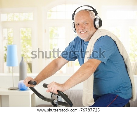 Active pensioner doing spinning on bike at home while listening to music, smiling at camera.?