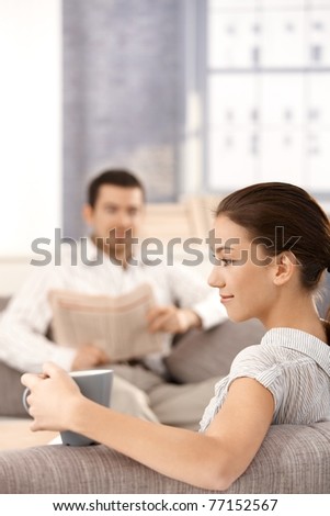 Young couple sitting in living room on sofa, man reading newspaper, woman drinking tea, smiling in front.?