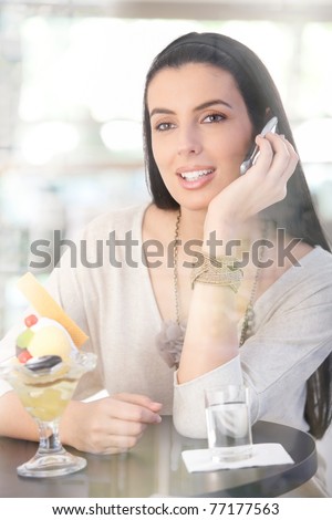 Happy woman sitting in cafe, having ice cream, talking on mobile phone, picture through window glass.?