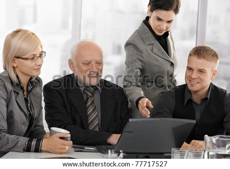 Businesspeople at meeting in office looking at laptop computer, businesswoman pointing at screen, smiling.?