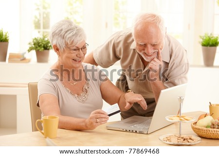 Happy older couple doing online shopping, laughing wife pointing at screen of laptop computer.?