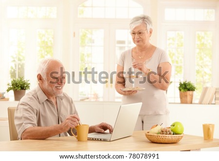 Happy senior man using laptop computer and having coffee at kitchen table, wife serving cookies.?
