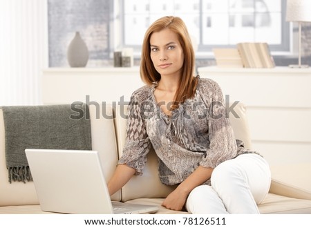 Portrait of attractive woman sitting on couch with laptop computer, looking at camera, smiling.?
