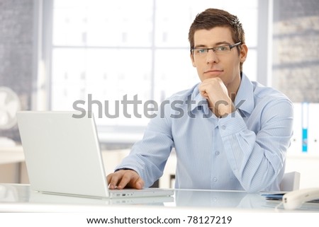 Portrait of office worker man sitting at office desk using laptop computer, looking at camera.?