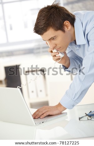 Young office worker man speaking on landline phone using laptop computer, looking at screen.?