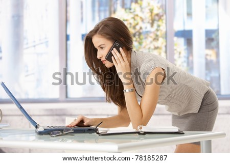 Pretty businesswoman concentrating on multitasking at desk, using laptop computer, talking on mobile phone.?