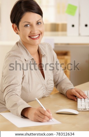 Happy office worker working at desk writing notes. Looking at camera, smiling.?