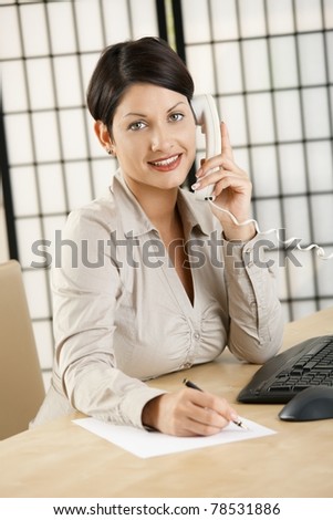 Young businesswoman sitting at desk in office, talking on phone, writing notes, smiling.?