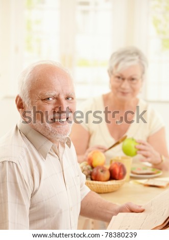 Portrait of smiling older man at kitchen table having breakfast, holding newspaper, with wife in background.?