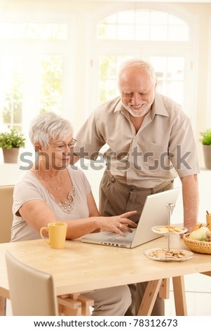 Smiling elderly couple using laptop computer in dining room, smiling, wife pointing at screen.?