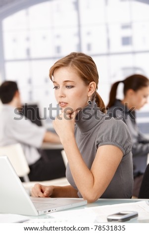 Young female office worker sitting at desk in office working on laptop, colleagues working in the background.?