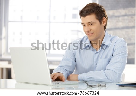 Young businessman working in office, sitting at desk, looking at laptop computer screen, smiling.?