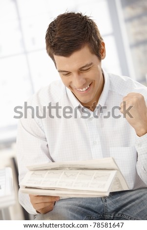 Young businessman happy about news reading in papers, raising fist, laughing.?