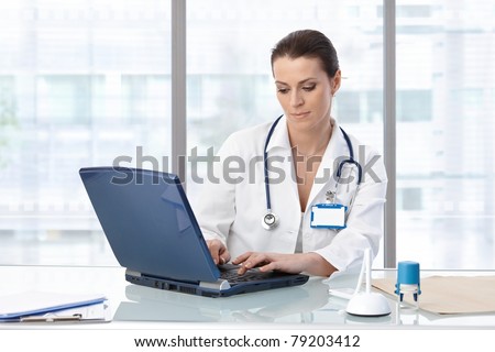 Female doctor sitting at table with laptop, looking down, working.?