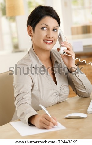 Happy businesswoman working at desk in office, writing notes while talking on phone