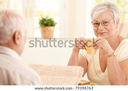 Portrait of senior woman having morning coffee, smiling at camera, sitting in kitchen with husband.?