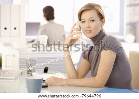 Young businesswoman sitting at office desk, talking on landline phone. looking at camera.?