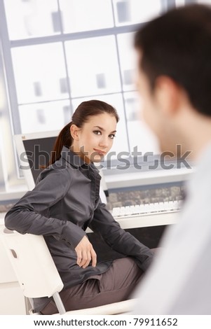 Young attractive businesswoman sitting at desk, turning back, smiling at her colleague.?