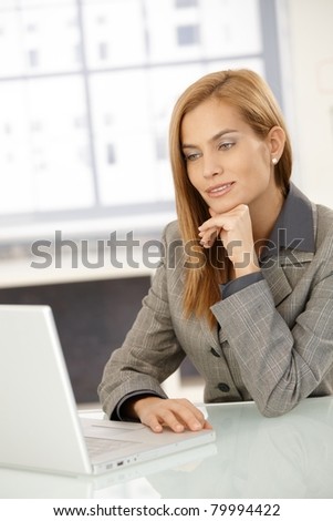 Smart businesswoman sitting in office, working on laptop computer, looking at screen, smiling.?