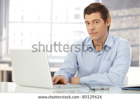 Portrait of young professional in office typing on laptop computer, looking at camera, smiling.?