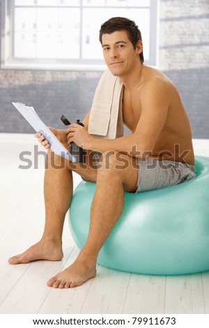 Handsome guy sitting on fit ball in gym in break of training, checking clipboard and drinking sports drink.?