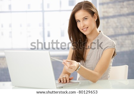 Cheerful young woman pointing at screen while reading on laptop computer, smiling at camera.?