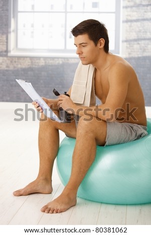 Young man sitting on gym ball in break of training, drinking sports drink and checking training program on clipboard.?