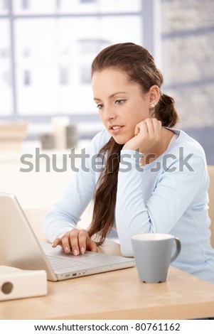 Pretty girl working at home, using laptop, looking at screen, sitting by table.?