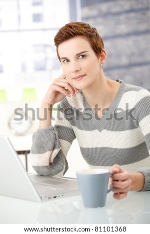 Portrait of young woman sitting at table with coffee mug and laptop computer, smiling at camera.?