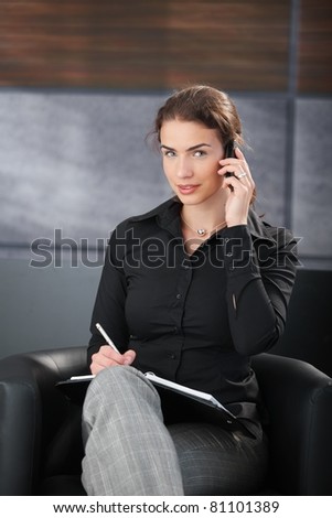 Attractive businesswoman chatting on mobile, writing notes, smiling in trendy office.?