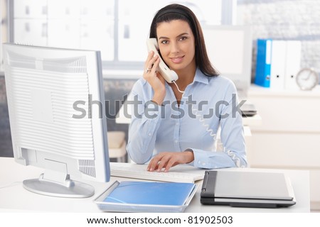 Portrait of smiling woman working in office, using computer and landline phone.?